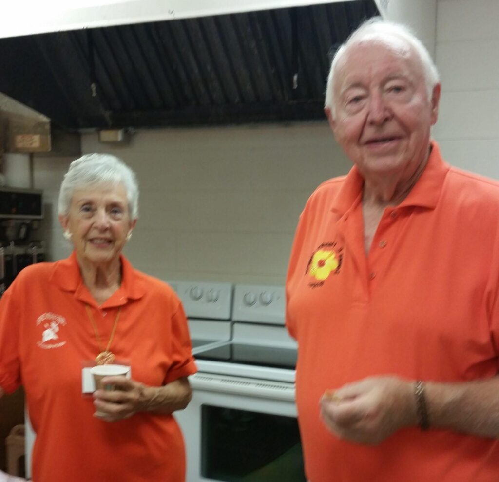 Two elderly people smiling in an indoor setting, wearing matching orange shirts.