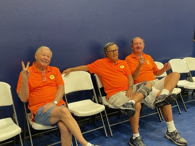 Three elderly men in orange shirts sitting on chairs outdoors.
